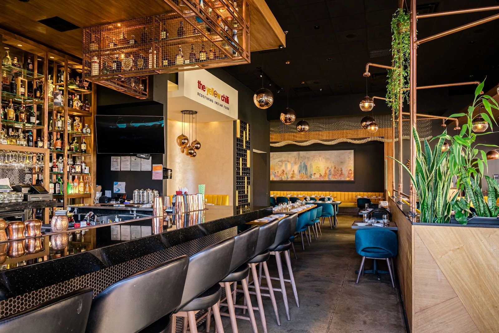 Modern cocktail bar area at The Yellow Chilli USA featuring dark marble counter, black barstools, illuminated liquor shelves, and copper accents by Urban Box Studio