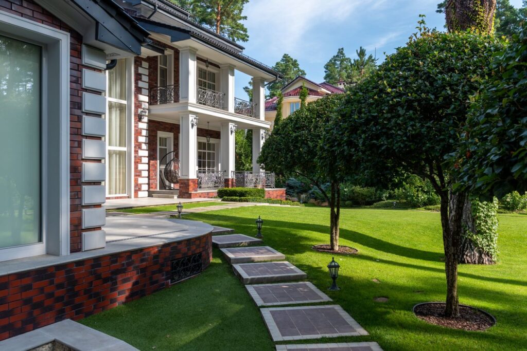 Modern house near backyard with path near green grass and plants with trees under blue sky in summer sunny day